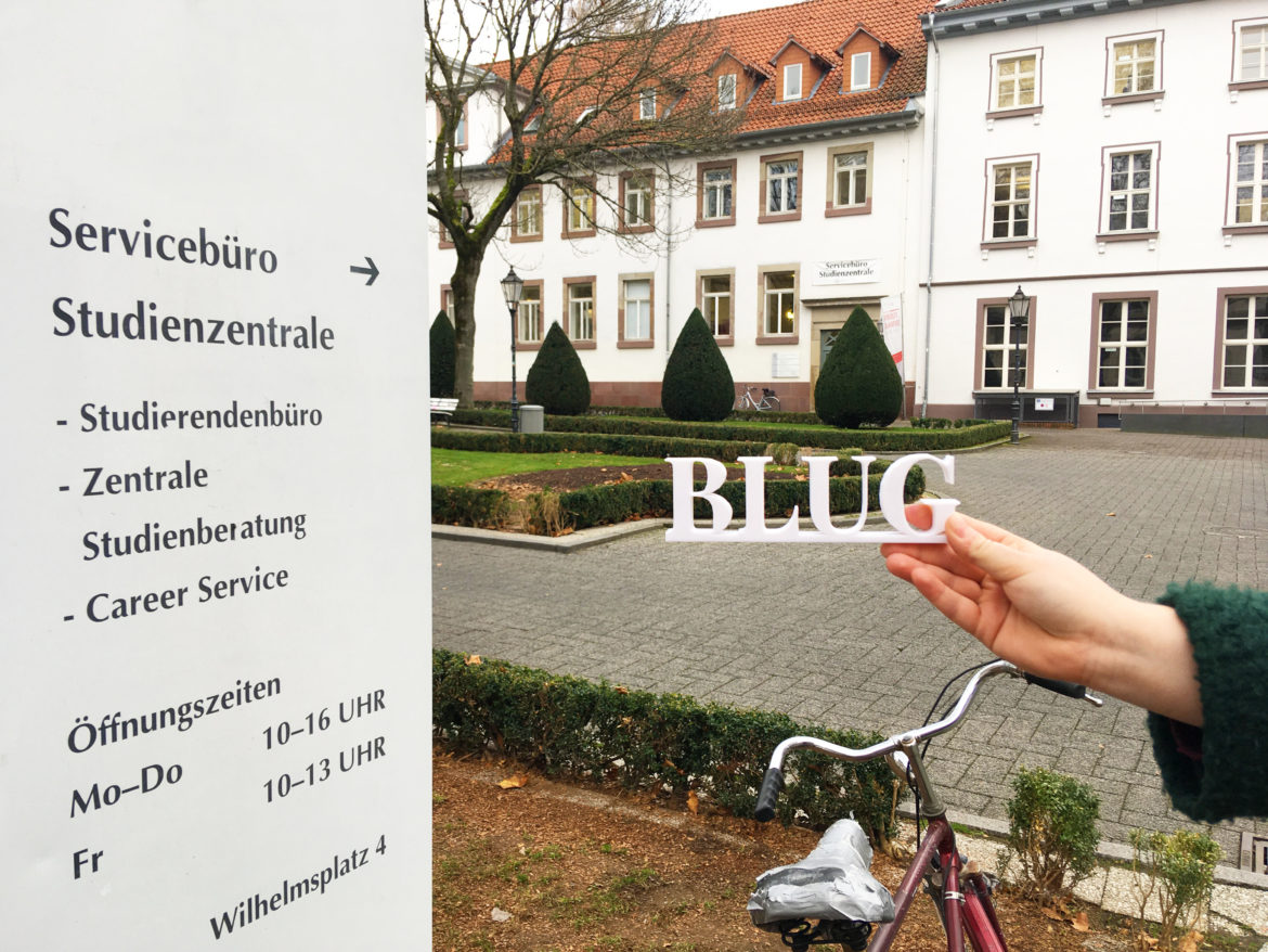 The BLUG Logo in front of a "Office of Student Affairs" sign with the actual office and the Wilhelmsplatz in the background