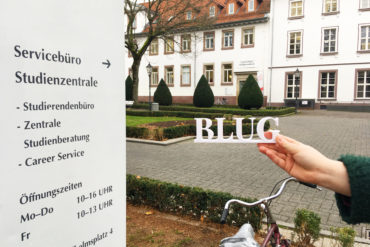 The BLUG Logo in front of a "Office of Student Affairs" sign with the actual office and the Wilhelmsplatz in the background